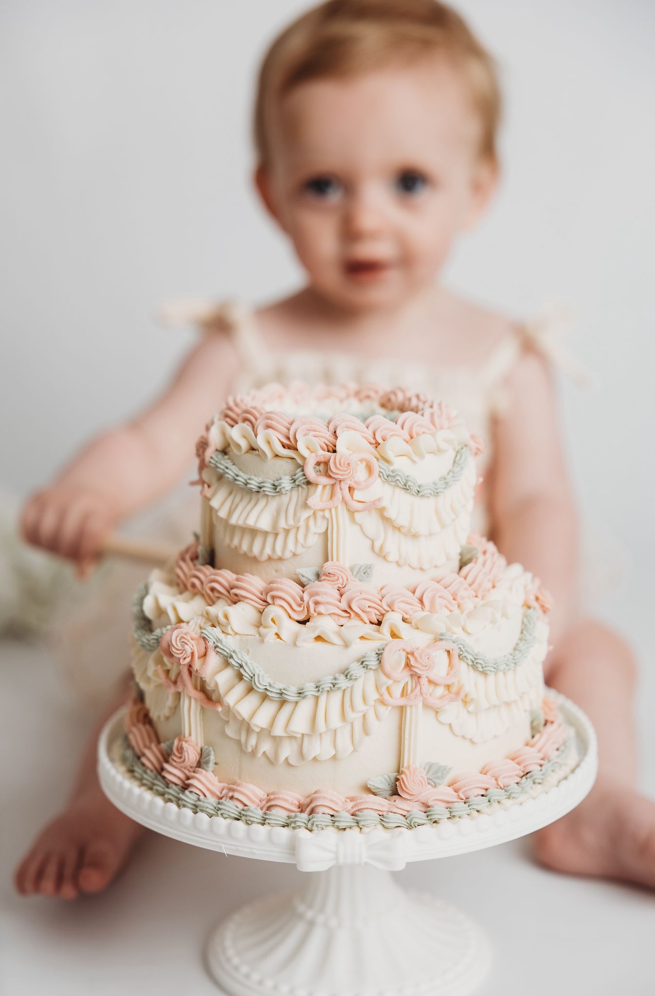 Pink and ivory two-tier birthday cake with delicate buttercream piping, photographed with a child celebrating their first birthday