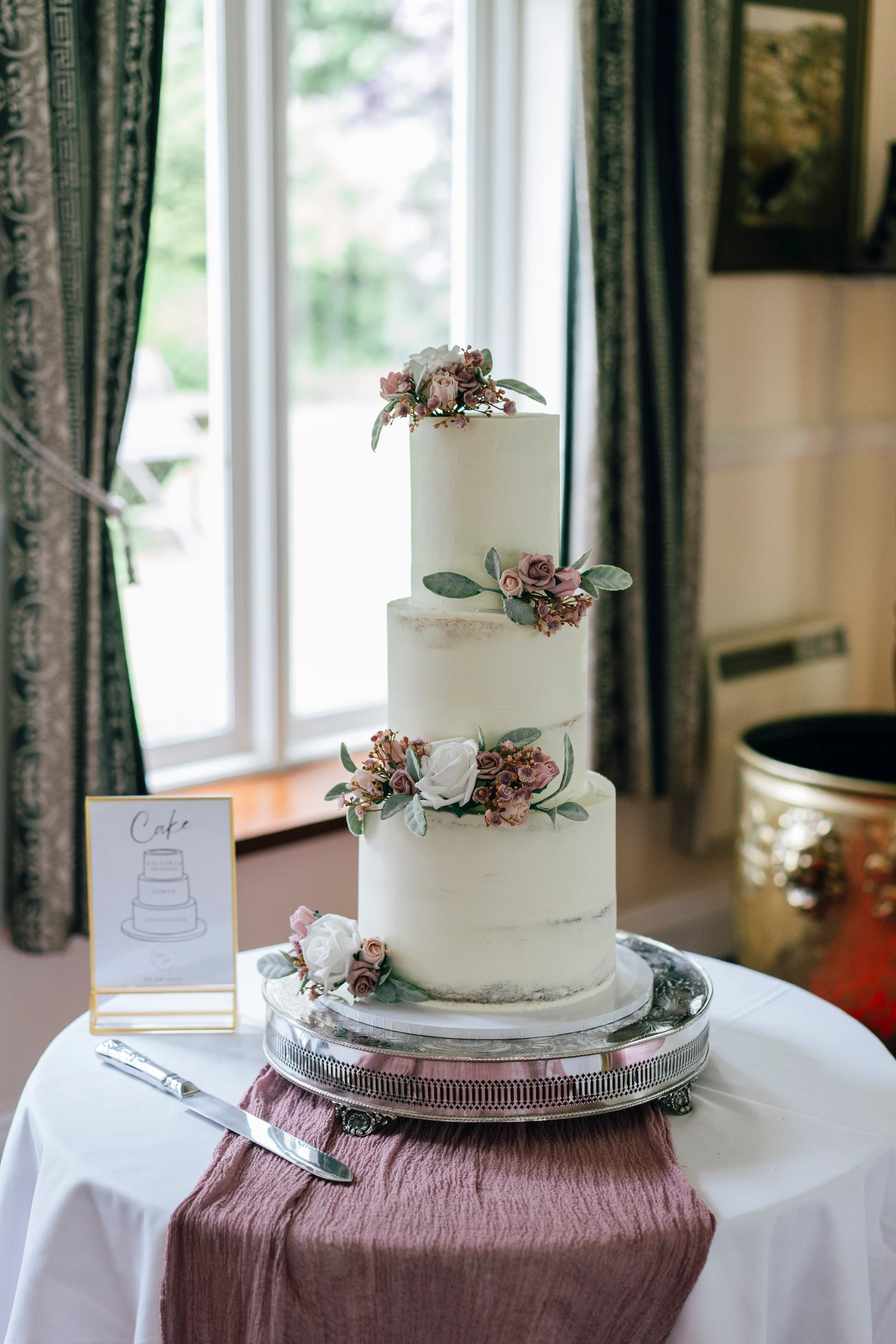 Rustic three-tier wedding cake with pink and ivory flowers on a silver stand beside a window